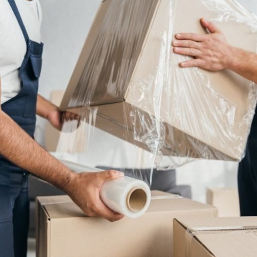 cropped view of movers in overalls wrapping box with stretch film